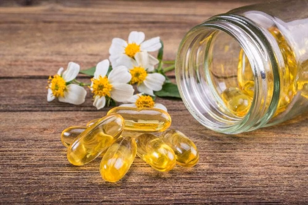 Các viên nang vitamin E trong lọ thủy tinh đặt cạnh hoa cúc trên bàn gỗ
