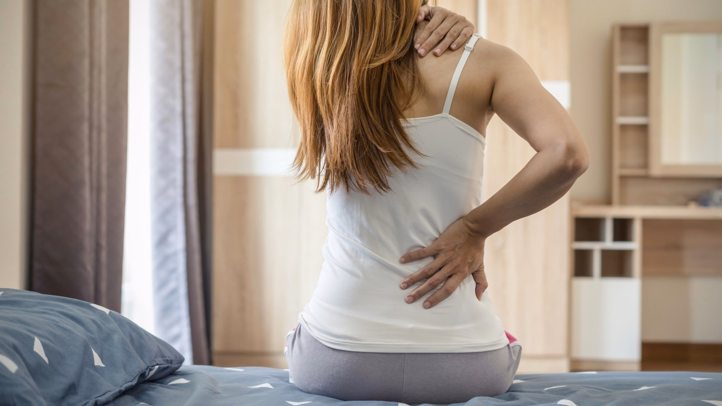 Người phụ nữ ngồi trên giường bị đau mỏi vùng cổ vai gáy và đau nhức lưng